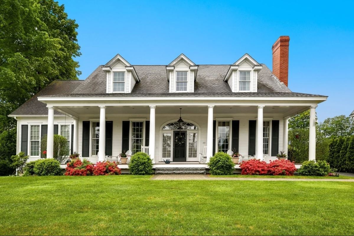 Cette maison de Cape Cod est blanche avec des volets noirs et une cheminée centrale comprend des piliers et un porche.